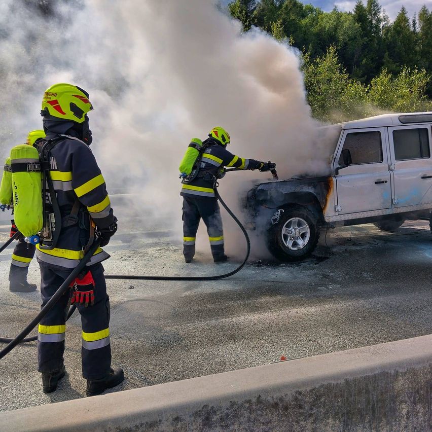 Feuerwehrleute verwenden einen Schlauch, um ein brennendes Auto auf der Straße zu löschen, umgeben von Rauch. Bäume und eine Betonbarriere sind im Hintergrund.