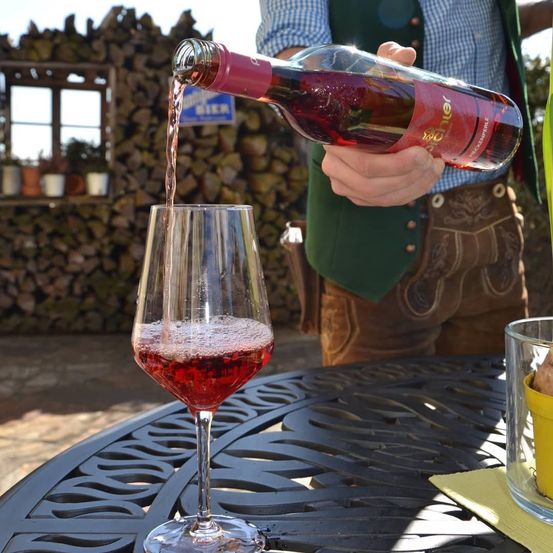 Ein Mann gießt roten Wein in ein Glas auf einem Metalltisch im Freien und trägt traditionelle deutsche Kleidung.