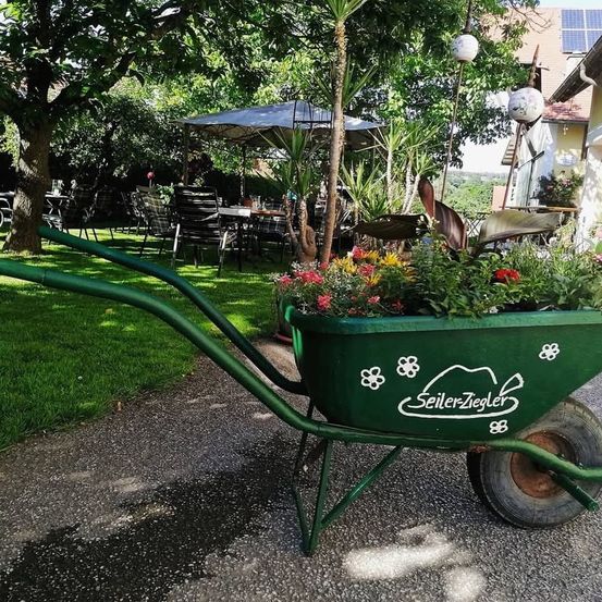 Ein grüner Schubkarren mit Blumen steht auf einem Kiesweg in einem Garten. Bäume, Pflanzen und ein Tisch mit Stühlen sind im Hintergrund.