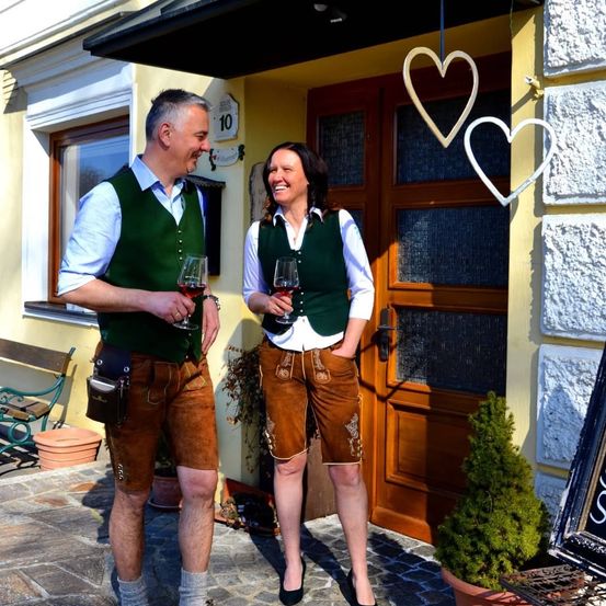 Ein Mann und eine Frau in traditioneller Kleidung stehen vor einem Gebäude, lächeln und halten Weinläser in der Hand. Sie stehen vor einer Holztür mit Herzdekorationen darüber.