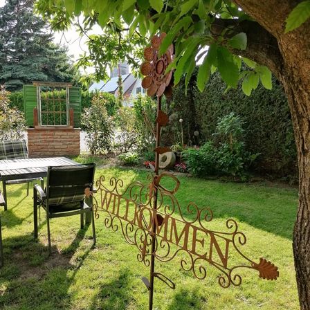 Ein Garten mit Tisch, Stühlen und einem 'Willkommen'-Schild. Es gibt Pflanzen, eine Ziegelmauer und ein Haus in der Ferne.