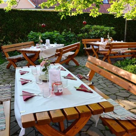 Ein Außenbereich mit hölzernen Bänken und Tischen, wo ein Tisch mit einer weißen Tischdecke und ein anderer Tisch mit Servietten und Gläsern gedeckt ist.