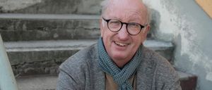 Ein älterer Mann mit Brille, Schal und Mantel sitzt auf Betonstufen, lächelt und schaut zur Kamera.