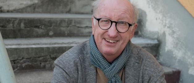 Ein älterer Mann mit Brille, Schal und Mantel sitzt auf Betonstufen, lächelt und schaut zur Kamera.