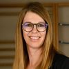 Eine Frau mit blonden Haaren, die eine Brille und einen schwarzen Blazer trägt, lächelt vor einer Holzstruktur.
