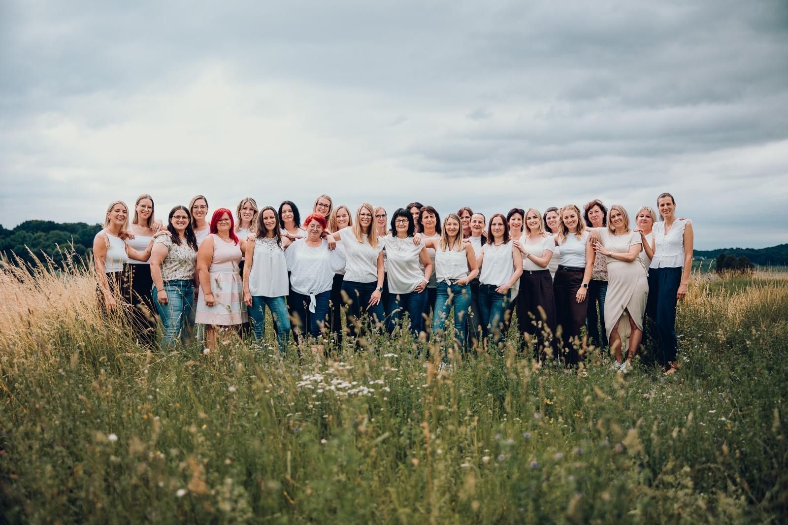 Eine große Gruppe von Frauen in weißen Kleidern und Jeans posiert für ein Foto in einem Grasfeld unter einem bewölkten Himmel.