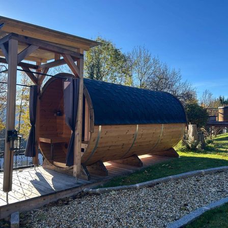 Eine Holzsauna steht auf einer Holzterrasse, umgeben von einer Holzkonstruktion. Die Terrasse ist von Kies umgeben. Dahinter befindet sich eine Rasenfläche mit Bäumen.