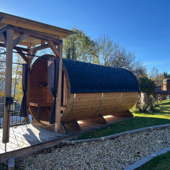 Eine Holzsauna steht auf einer Holzterrasse, umgeben von einer Holzkonstruktion. Die Terrasse ist von Kies umgeben. Dahinter befindet sich eine Rasenfläche mit Bäumen.