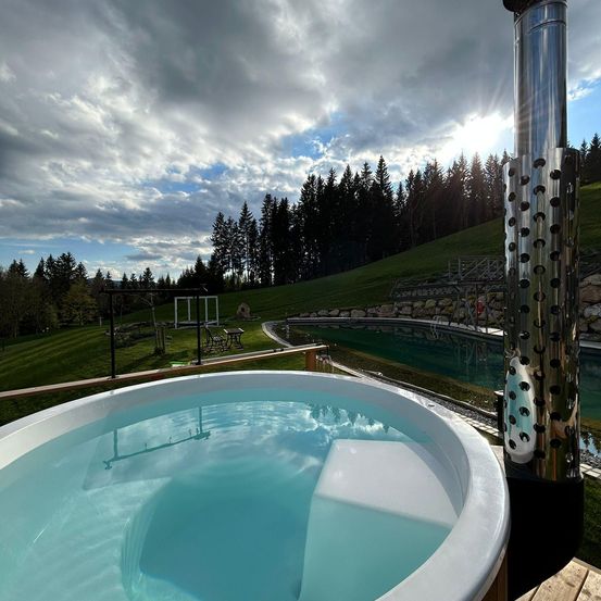 Ein großer Außen-Whirlpool mit klarem Wasser, auf einer Holzterrasse mit Blick auf einen grünen Hügel und Bäume.