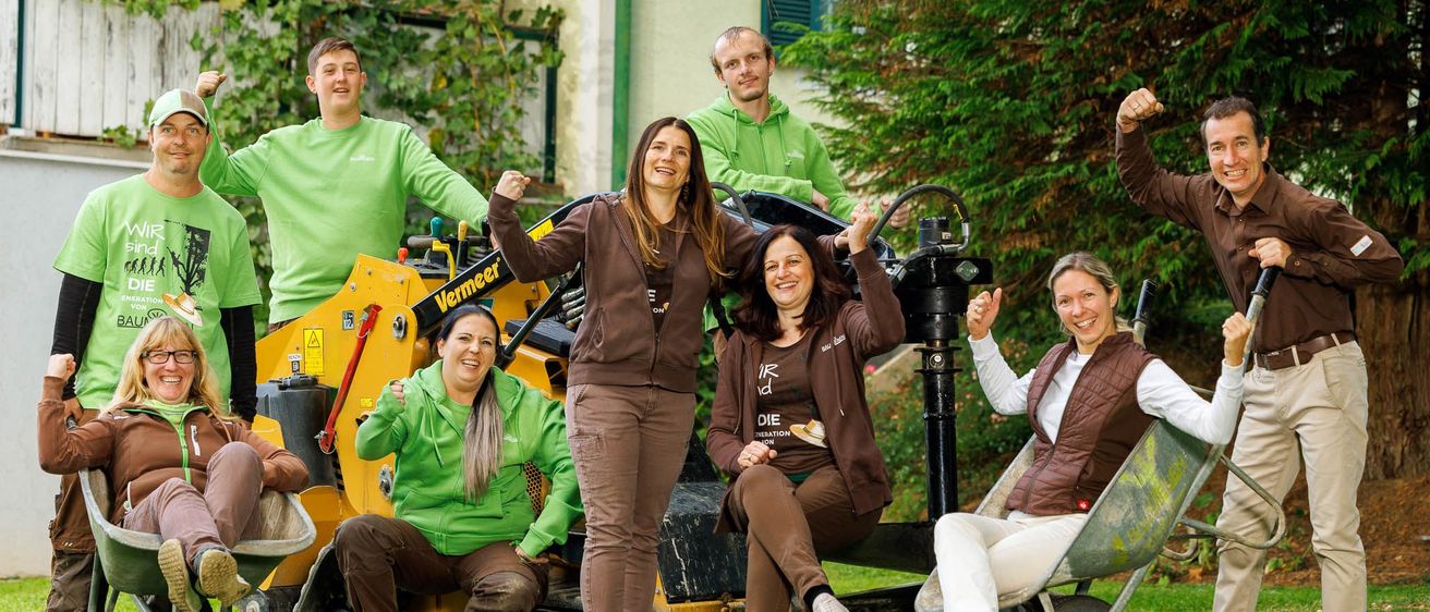Eine Gruppe von Menschen in grünen und braunen Uniformen, die um eine gelbe Maschine herum sitzen und stehen, posiert für ein Foto auf einem Rasenfeld mit einem Gebäude im Hintergrund.
