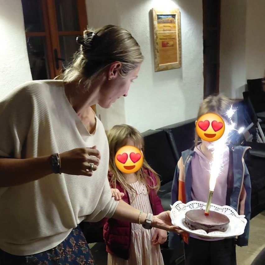 Eine Frau steht und hält einen Kuchen, während zwei Kinder mit Geburtstagsfunken ins Gesicht schauen. Das Mädchen hat einen Herz-Emoji-Aufkleber im Gesicht.