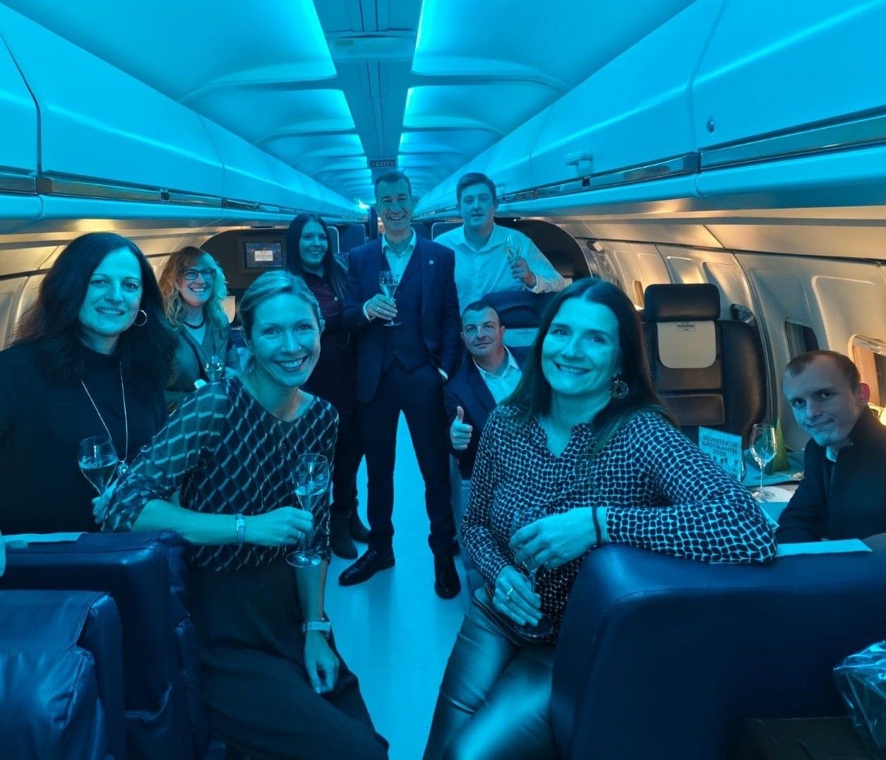 Eine Gruppe lächelnder Erwachsener in formeller Kleidung in einem Flugzeug, einige stehend und andere sitzend, halten Weingläser in der Hand, unter blauer Beleuchtung.