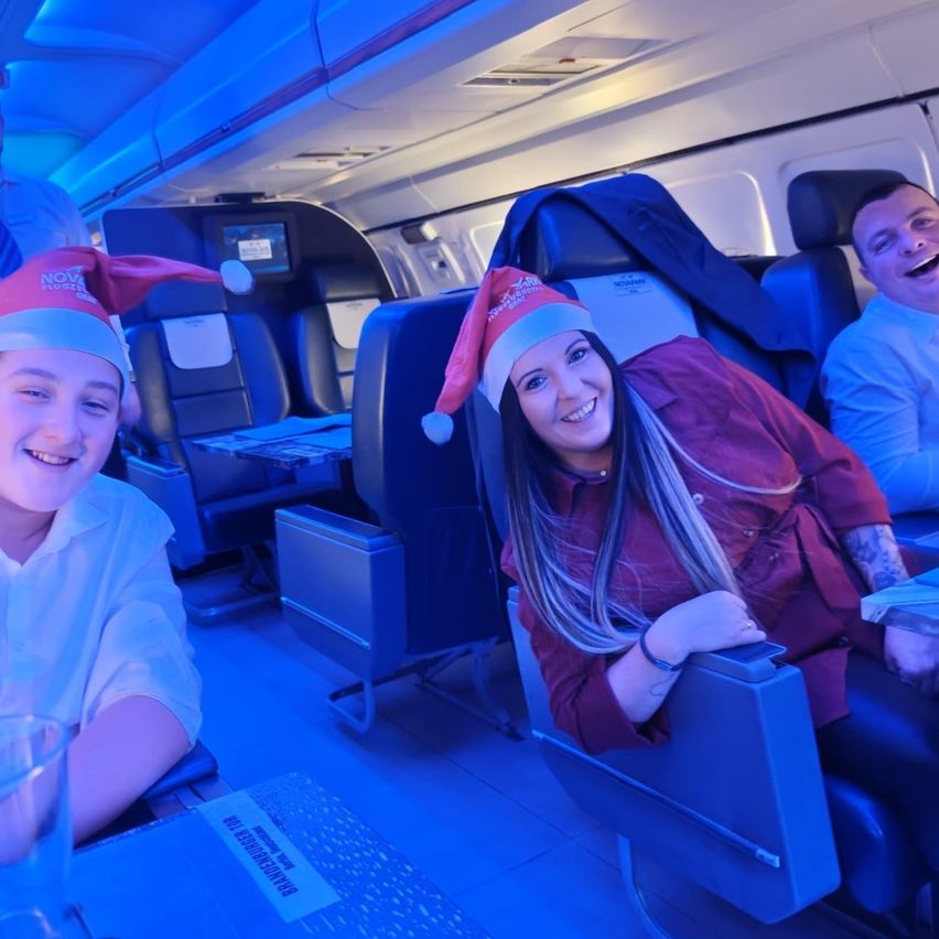 Eine Gruppe von Menschen sitzt in einem Flugzeug. Zwei Personen, ein Junge und eine Frau, tragen Weihnachtsmützen. Die Frau lächelt und posiert für ein Foto.