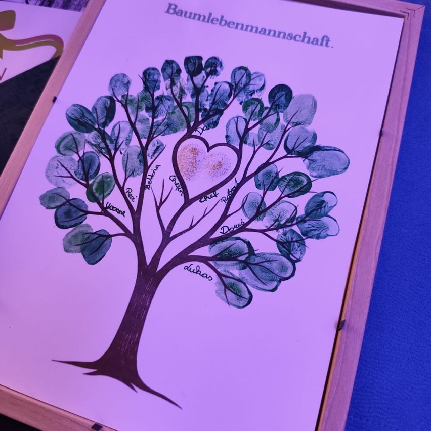 Ein gerahmtes Poster mit einem handgezeichneten Baum mit einem Herz in der Mitte, beschriftet mit 'Baumlebenmannschaft', mit Namen auf grünen Blättern.