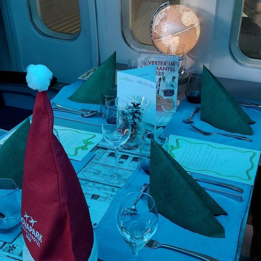 Ein Tisch ist für eine Feier gedeckt mit Weingläsern, Besteck und grünen Servietten. Ein roter Hut mit einer weißen Quaste liegt auf dem Tisch. Ein Globus und ein Buch mit dem Titel 'Silvester im Mantel 2025' stehen auf dem Tisch, und ein Fenster spiegelt den Globus wider.