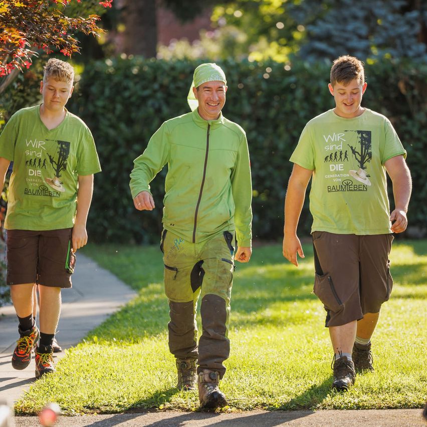 Drei Personen in grünen Shirts und Shorts laufen auf einem Rasen. Einer trägt einen grünen Hut, ein anderer hat eine grüne Jacke an. Sie alle lächeln.