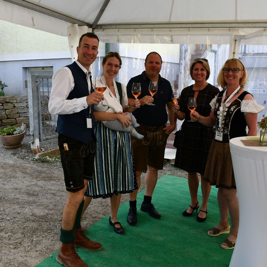 Five adults dressed in traditional Bavarian clothing stand under a white tent and hold wine glasses. Two women carry babies.