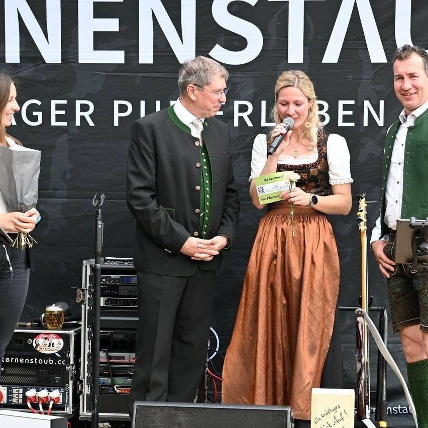 Drei Personen stehen auf der Bühne, eine Frau hält ein Mikrofon. Ein Mann im Anzug steht hinter ihr, und ein weiterer Mann steht zu ihrer rechten. Sie stehen vor einem Banner, das 'Pierler Ben' besagt.