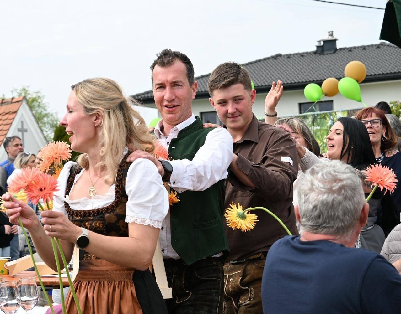Zwei Männer und eine Frau in traditioneller deutscher Kleidung stehen unter einer Gruppe von Menschen, halten Blumen und lächeln in die Kamera. Dahinter ist ein Haus mit grünen Luftballons und einem Schornstein zu sehen.