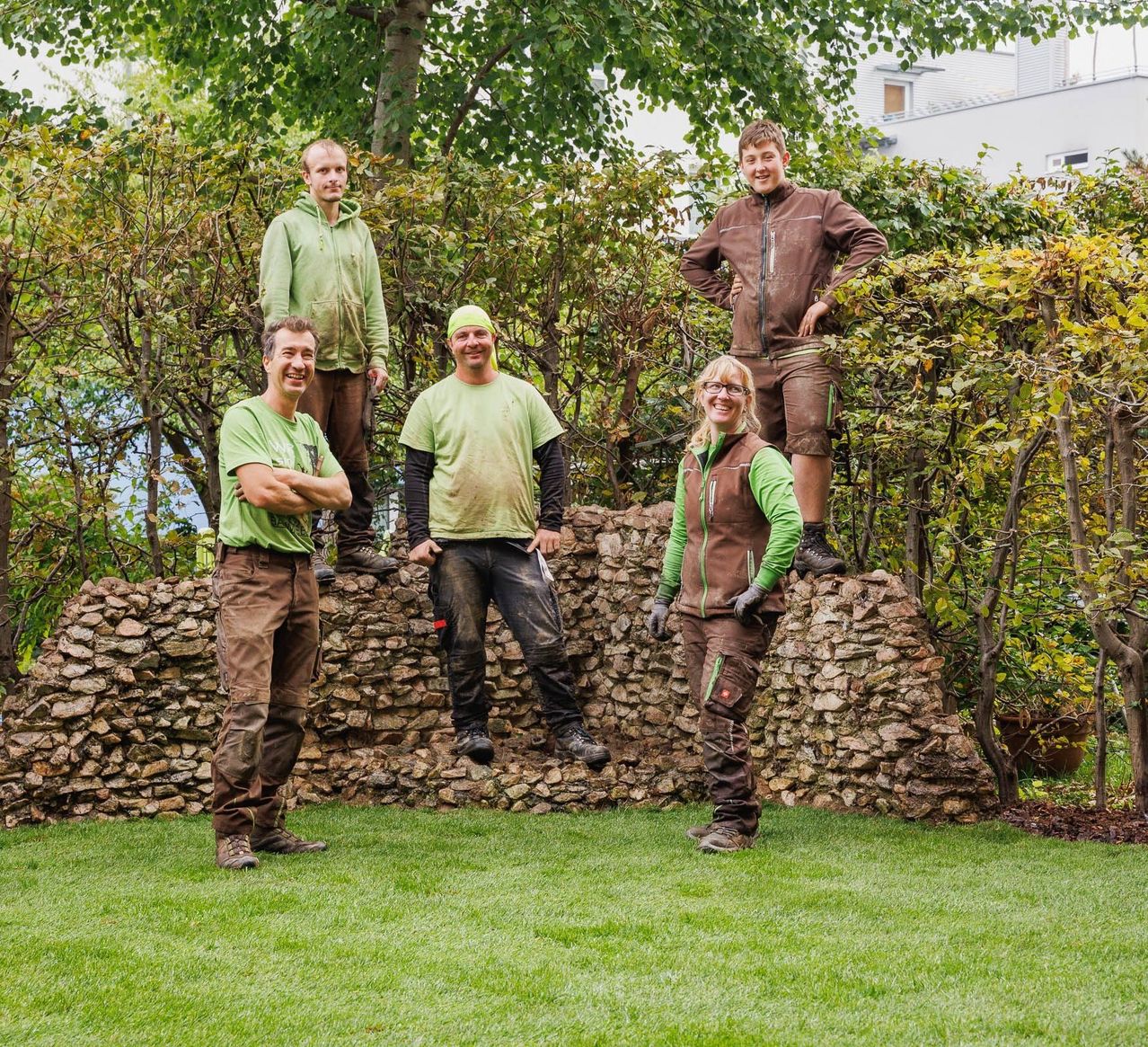 Fünf Menschen, alle in grüner oder brauner Arbeitskleidung, stehen auf einer Steinmauer. Sie scheinen für ein Foto zu posieren, mit einem Grasbereich und Pflanzen hinter ihnen.