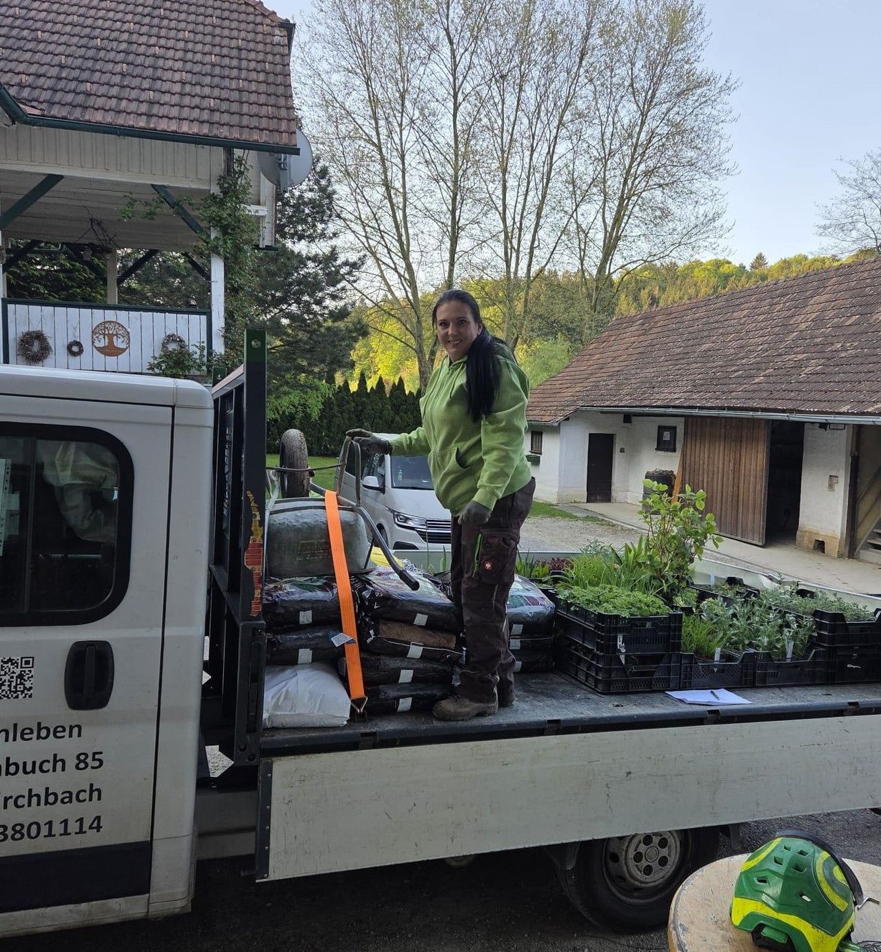 A woman in a green jacket is loading potted plants into the back of a truck. The truck is parked in front of a house with a brown roof.