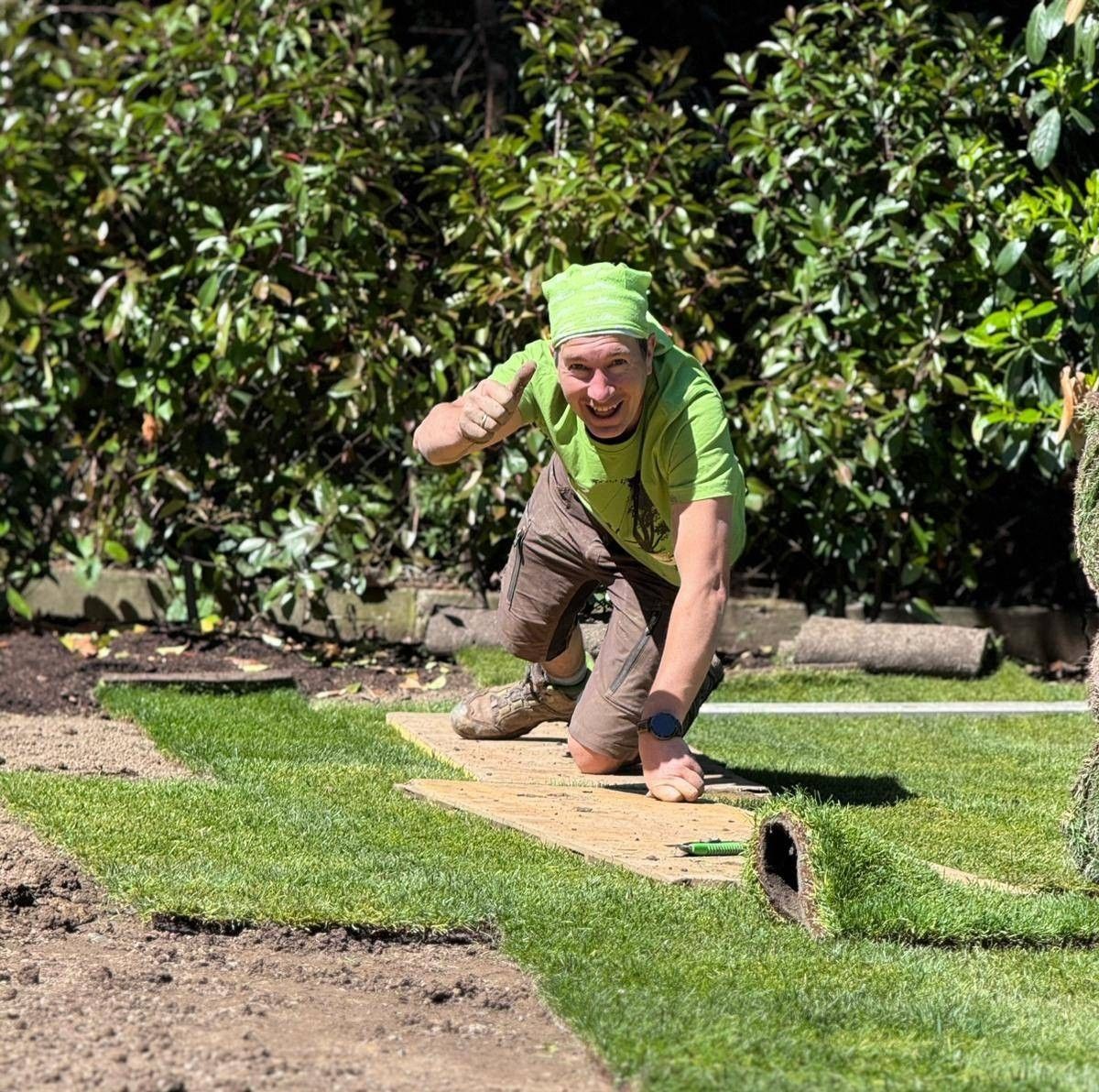 A man in green is installing artificial turf on a yard, smiling, and showing a thumbs up. He's on his knees with bushes and a wooden path behind him.