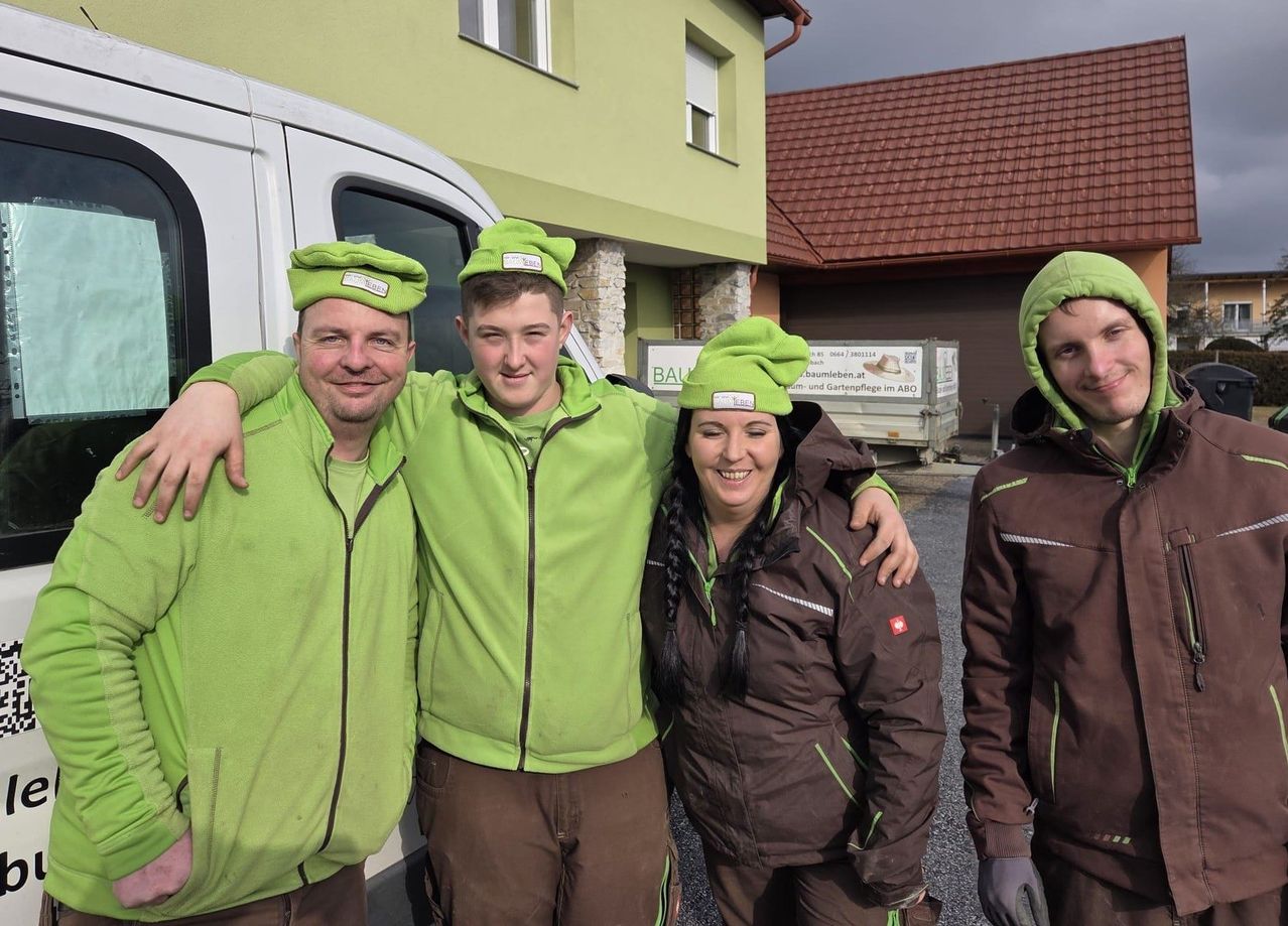 Vier Personen in grünen und braunen Jacken und Mützen stehen vor einem Haus. Sie lächeln.