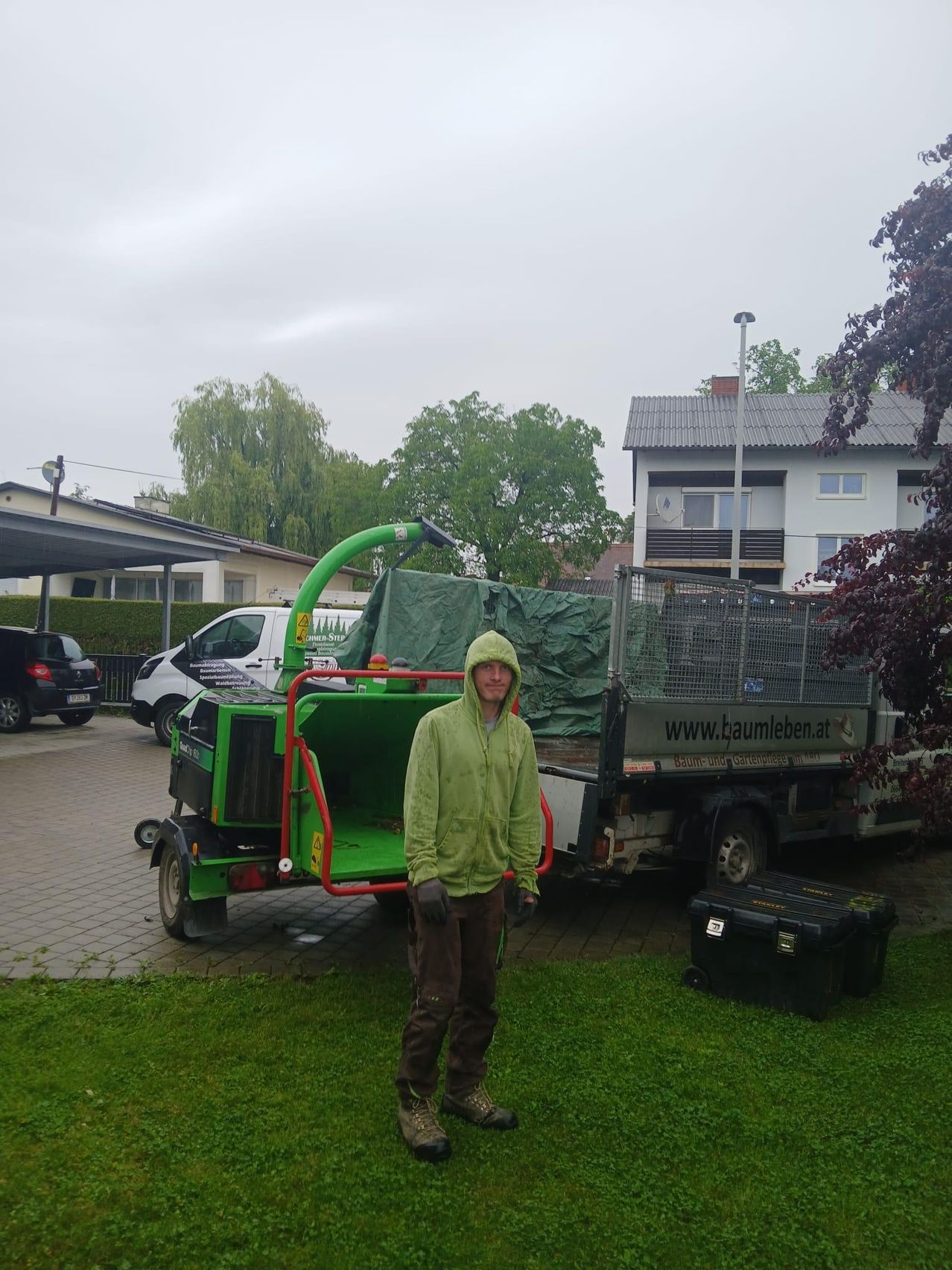 Ein Mann in einer grünen Jacke steht neben einem grünen Lkw mit einer Plane, der vor einem Haus geparkt ist.