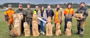 A group of people in jackets stand in a grassy field with carved owl sculptures. They are smiling and posing for a photo. The background shows trees and a cloudy sky.