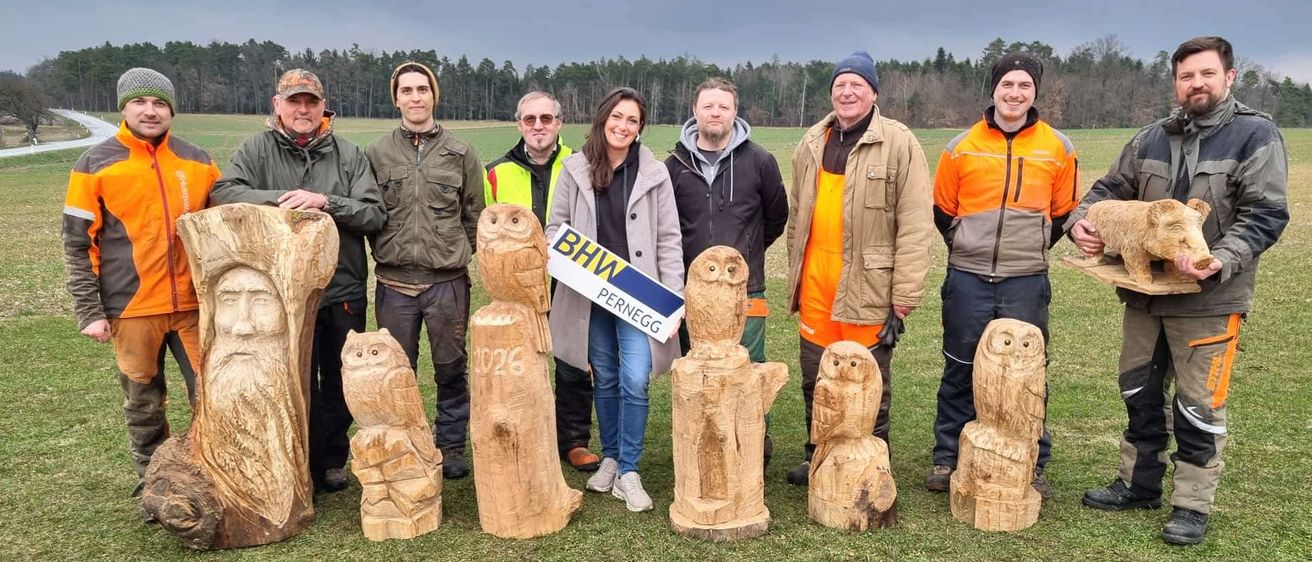 Eine Gruppe von Menschen in Jacken steht auf einem Grasfeld mit geschnitzten Eulen-Skulpturen. Sie lächeln und posieren für ein Foto. Im Hintergrund sind Bäume und ein bewölkter Himmel zu sehen.