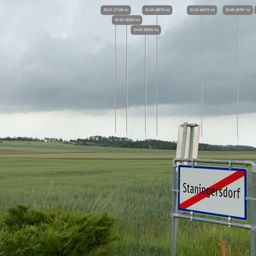 Eine ländliche Landschaft mit einem Schild, das Staningersdorf liest, unter einem bewölkten Himmel mit Windturbinen in der Ferne.