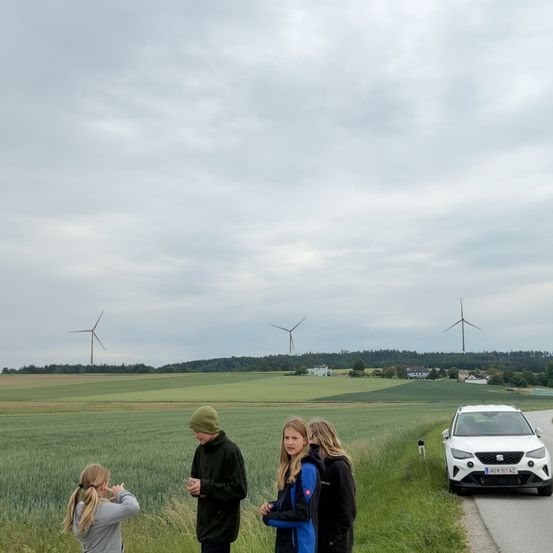 Drei Personen stehen in einem Feld mit hohem Gras. Ein weißes Auto ist am Straßenrand geparkt. In der Ferne sind Windturbinen und Bäume zu sehen. Der Himmel ist bewölkt.