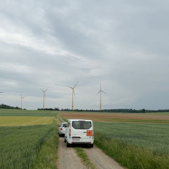 Ein weißer Van fährt mit einem Auto dahinter auf einem Feldweg durch ein Feld mit hohem Gras und Windturbinen im Hintergrund.
