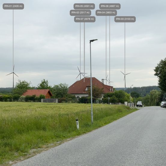 Eine Landschaft mit einer Straße und Windturbinen im Hintergrund, eine davon ist mit IRN-01 bei 2435 Metern gekennzeichnet. Ein Haus mit einem roten Dach ist in der Nähe.