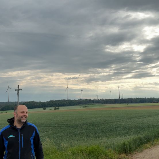 Ein Mann steht in einem Feld mit Windrädern in der Ferne unter einem bewölkten Himmel.