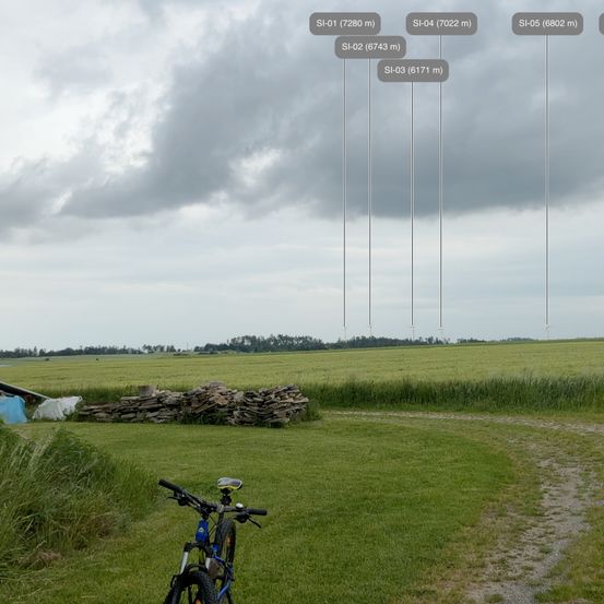 Ein Fahrrad ist auf einem Grasfeld geparkt. Links befindet sich ein Haufen Steine, im Hintergrund ein bewölkter Himmel. Das Bild enthält verschiedene Höhen eines Satelliten.