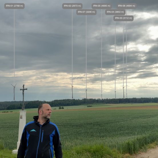 Ein Mann in einer blauen und schwarzen Jacke steht auf einem Feld und blickt auf Windturbinen in der Ferne. Der Himmel ist bewölkt und vor ihm steht ein hoher Kreuz.