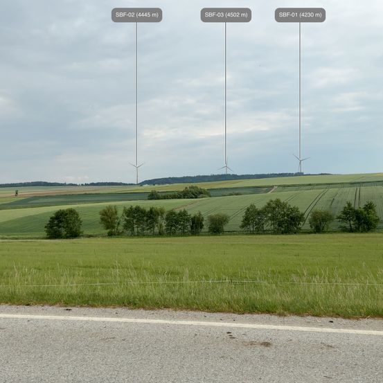 Eine ländliche Landschaft mit weiten Feldern und mehreren Windturbinen in der Ferne unter einem bewölkten Himmel.