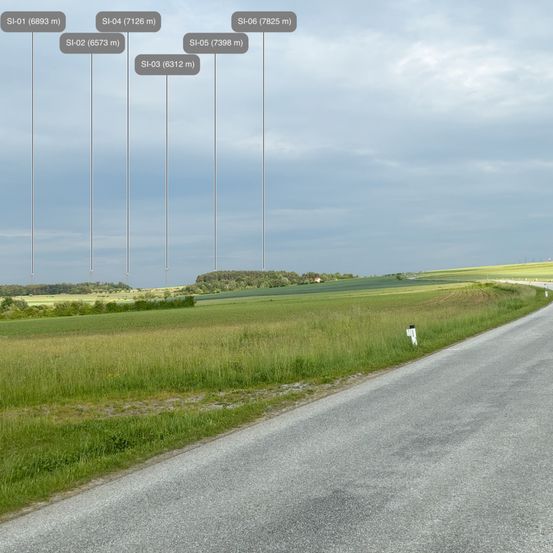 Eine ländliche Straße, umgeben von grünen Feldern. Am Himmel befinden sich sechs vertikale Linien mit Nummern und Höhen, die die Standorte und Höhen verschiedener Strukturen anzeigen.