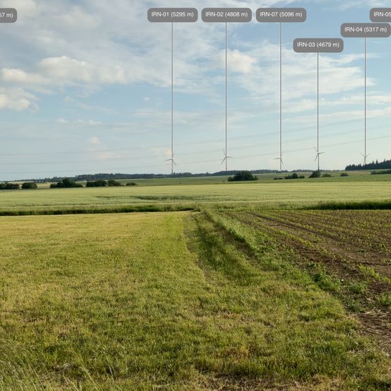 Eine Luftaufnahme eines Grasfeldes mit Windturbinen in der Ferne. Das Feld hat eine braune Stelle in der Mitte. Das Bild enthält mehrere Entfernungsmarkierungen.