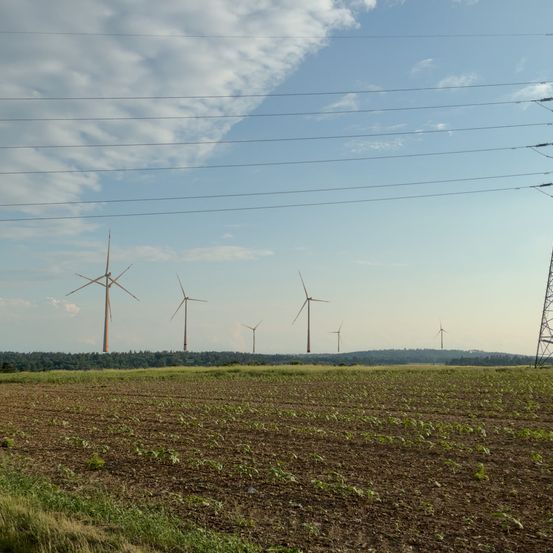 Eine Reihe von Windturbinen steht in einem weitläufigen, offenen Feld unter einem teilweise bewölkten Himmel. Die Turbinen sind in einer Reihe ausgerichtet, mit Stromleitungen darüber.
