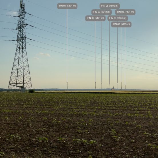 Ein großer Strommast steht in einem weiten Feld. Mehrere Stromleitungen erstrecken sich über die Landschaft, wobei Windturbinen in der Ferne sichtbar sind. Der Himmel ist klar und blau.