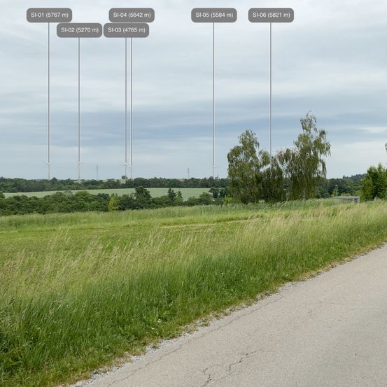 Ein weiter Blick auf ein saftiges Feld mit hohem Gras und einer Straße auf der rechten Seite. Bäume säumen den Horizont, und in der Ferne gibt es hohe Strukturen, die mit SI-01 bis SI-06 beschriftet sind. Der Himmel ist bewölkt.