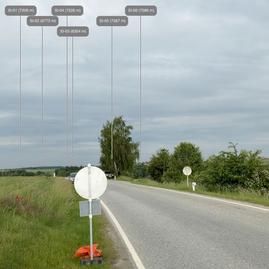 Eine Landstraße mit hohen Metallmasten, zwei Autos in der Ferne und einem Schild am Straßenrand.