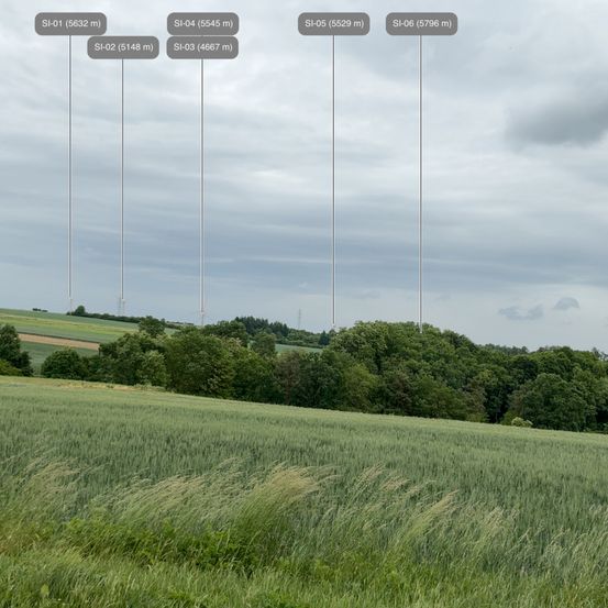 Ein ländliches Feld mit hohem Gras und Bäumen unter einem bewölkten Himmel. Mehrere Pfosten mit Nummern und Höhenangaben sind sichtbar.