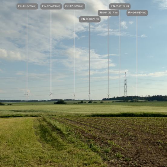 Eine Luftaufnahme einer ländlichen Landschaft mit grünen und braunen Feldern. Windräder und Stromleitungen sind sichtbar. Der Himmel ist teilweise bewölkt.