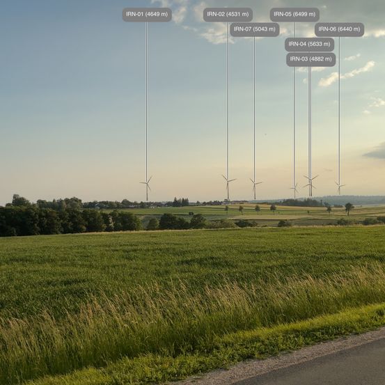 Offenes Feld mit mehreren Windturbinen, jede mit einer Kennzeichnungsnummer und Höhe. Der Himmel ist blau mit Wolken.