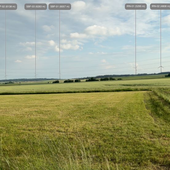 Ein weitläufiges Grasfeld unter einem teilweise bewölkten Himmel, mit mehreren Windturbinen in der Ferne. Das Feld erstreckt sich weit und wird von einem kleinen Pfad durchquert.