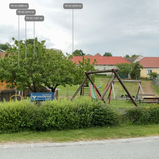 Ein kleiner Park mit Spielplatz wird gezeigt. Ein Volksbank-Schild befindet sich links. Ein Haus mit rotem Dach ist im Hintergrund.