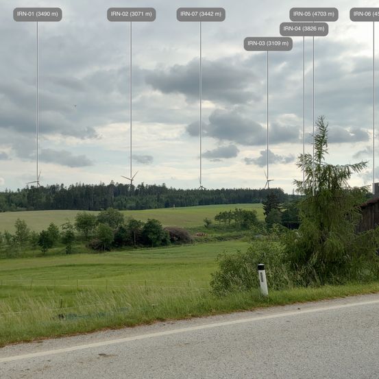 Eine ländliche Landschaft mit grünen Feldern und Bäumen. Mehrere Windturbinen sind in der Ferne sichtbar. Eine Straße mit einer weißen Linie erstreckt sich. Über der Landschaft sind mehrere Höhenangaben vorhanden.